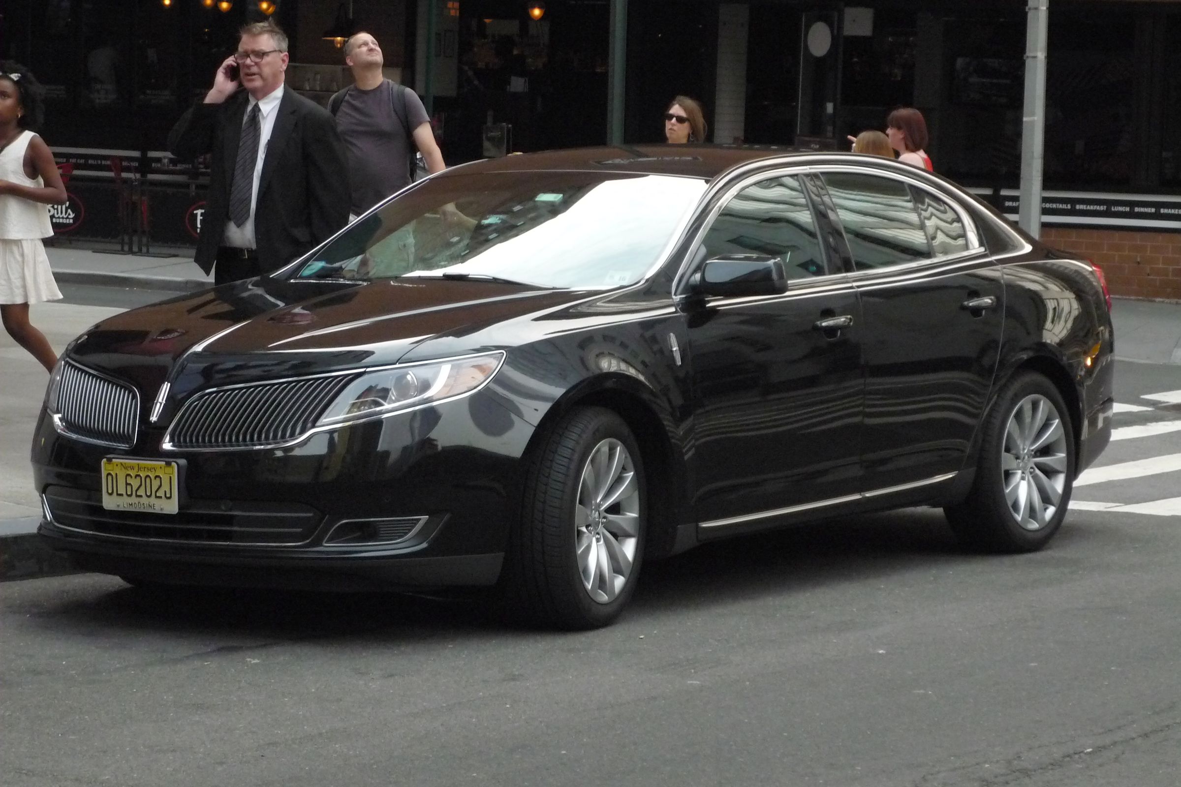 A black chauffeured sedan stopped at a kerb with a driver beside it.