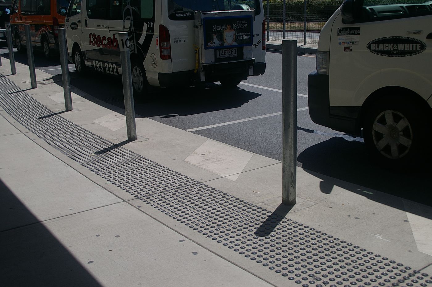 Shuttle vans queued along a covered airport kerb behind steel bollards.